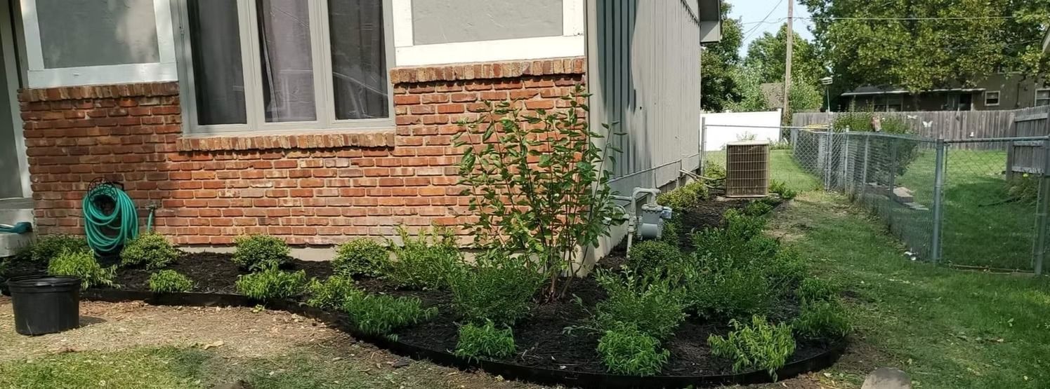 A house with brick facade, garden bed with shrubs, and a chain-link fence.