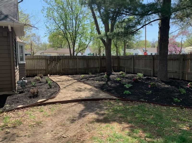 Backyard with gravel path, flower beds, trees, and wooden fence on a sunny day.