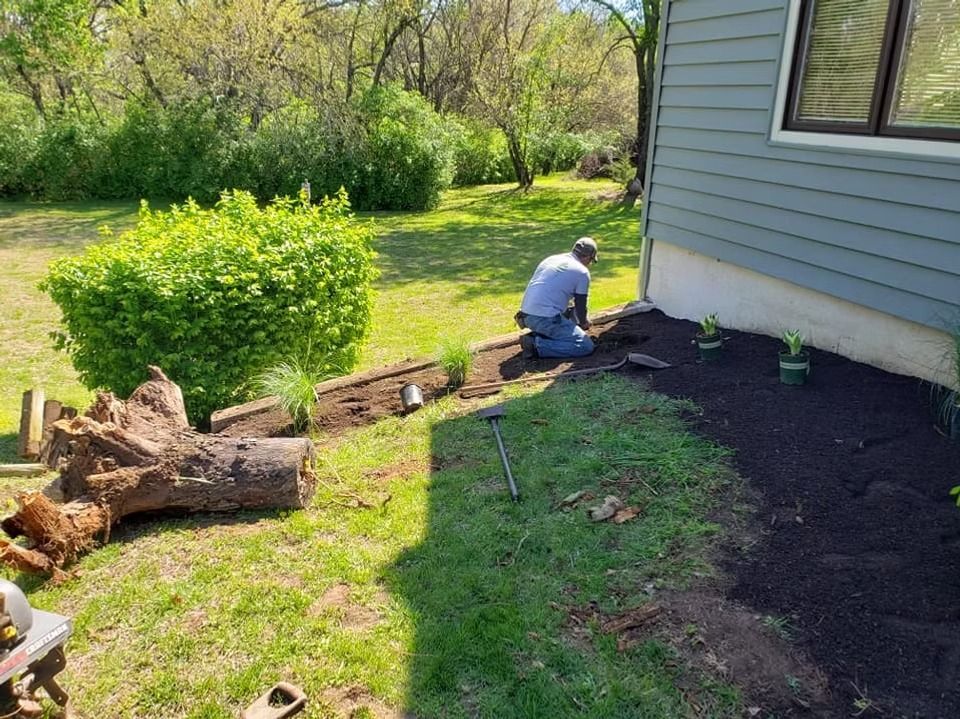 Man gardening next to house, planting in dark mulch, with green lawn and bush nearby.