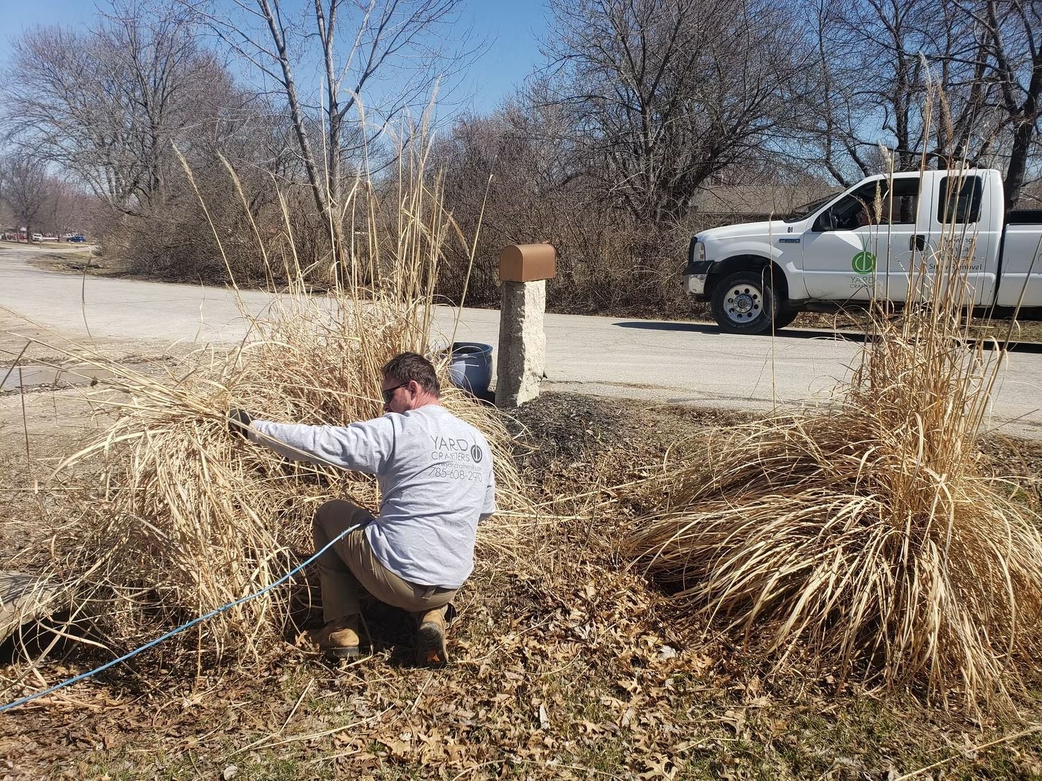 Man kneeling, trimming dry grasses along a roadside. A white truck is parked nearby. Sunny day.