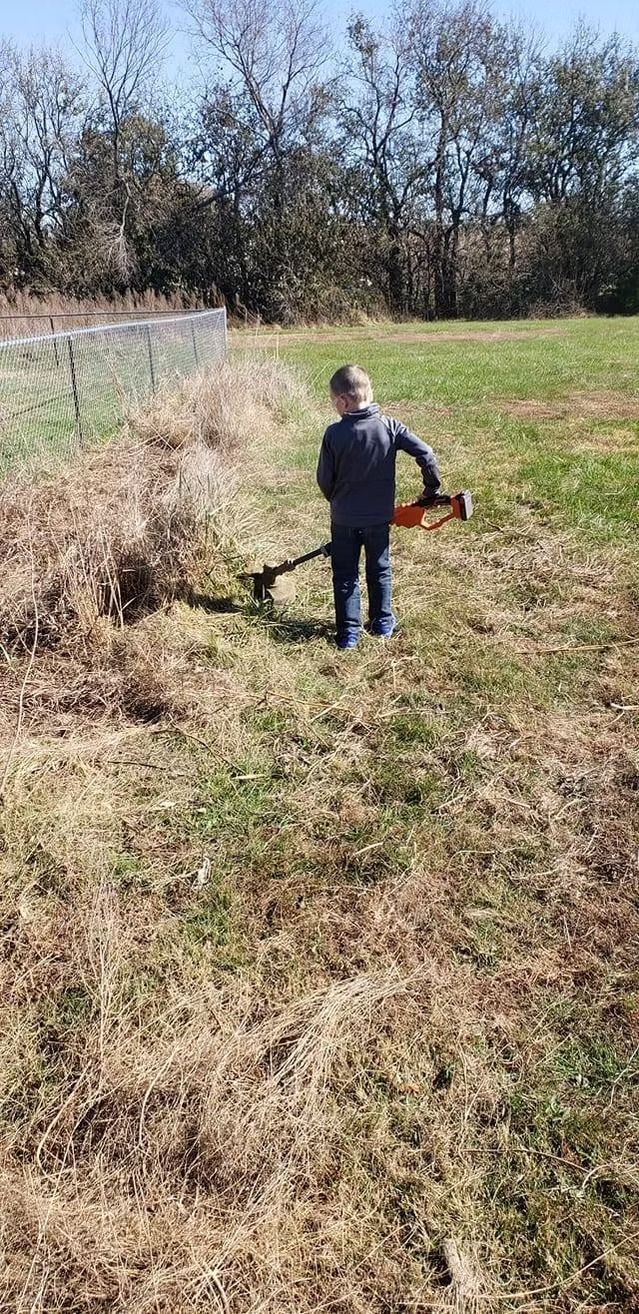 A child using a weed wacker in a grassy field, trees in the background.