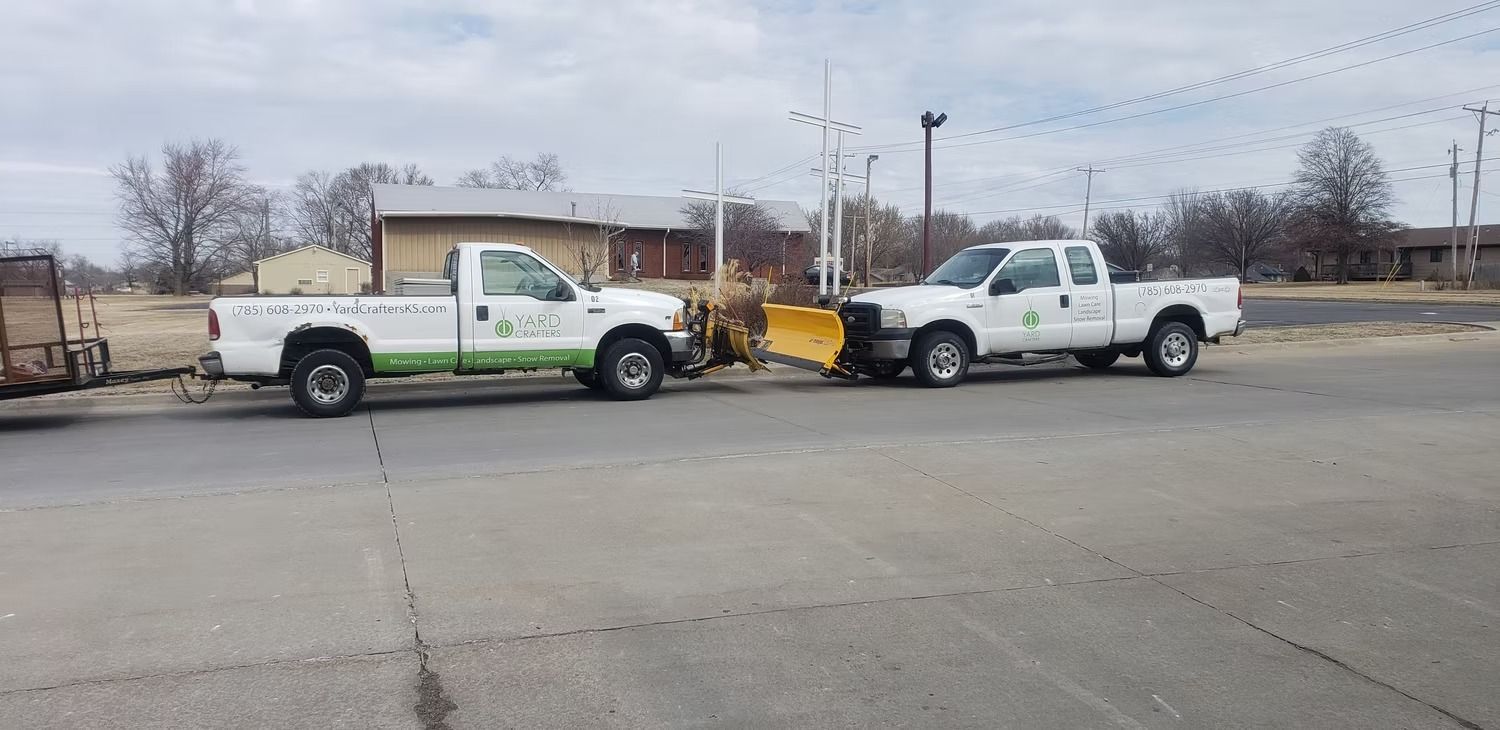 Two white trucks with snowplows parked on a gray asphalt surface. Buildings and trees in the background.