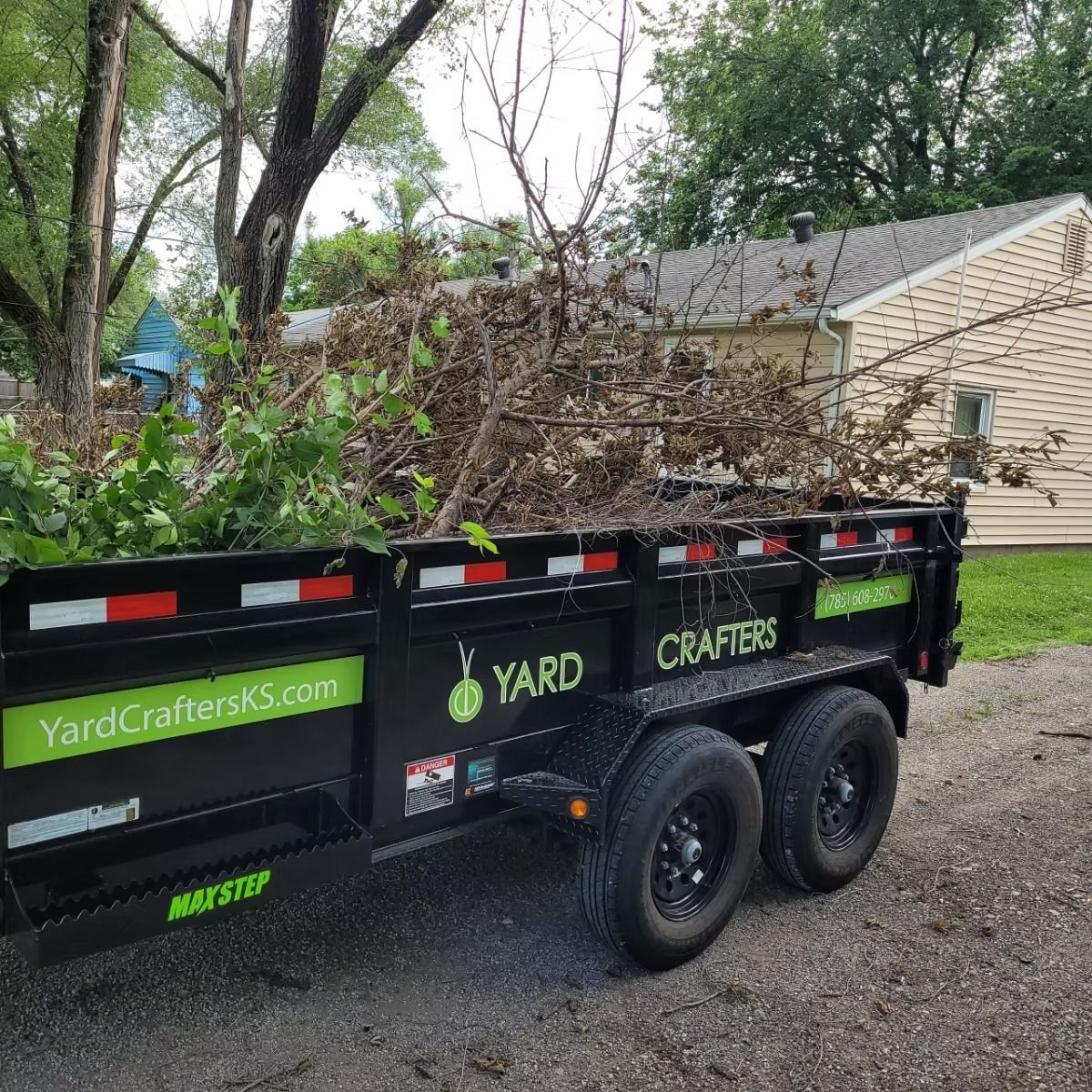 A black trailer filled with tree branches, parked in front of a house. 