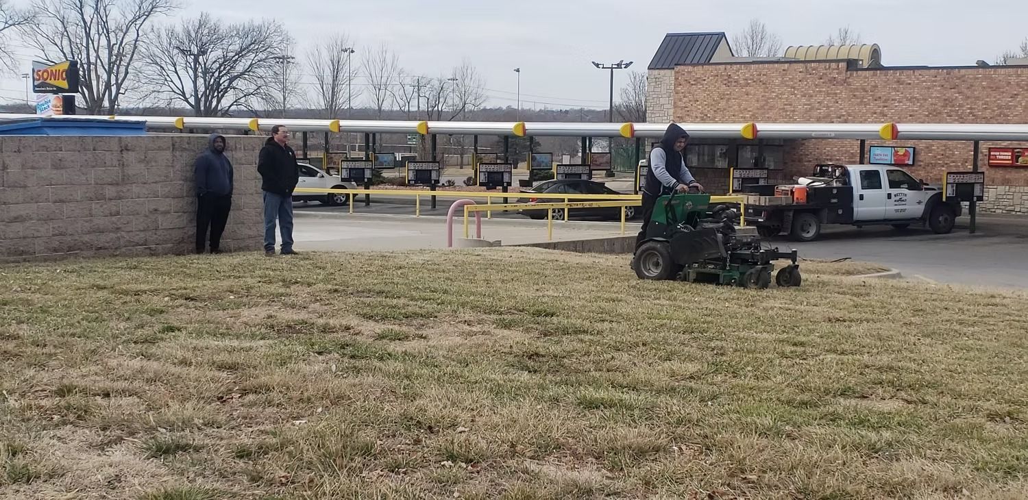 Man operating lawn aerator near a gas station with people, a truck, and a brick building.