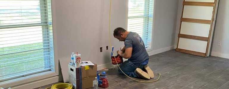 A man is kneeling down in a room working on a wall.