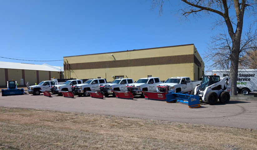 A line of white pickup trucks with snow plows attached and a skid steer parked in front of a tan commercial building.