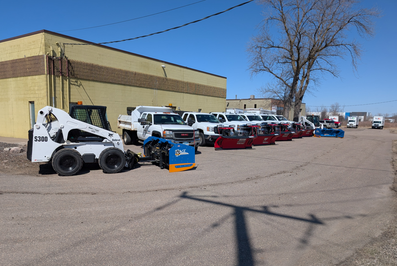 A row of parked white pickup trucks with snow plows attached and a white skid steer loader beside a yellow building.