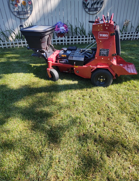 A red Toro stand-on spreader sprayer parked on a green lawn in front of a white fence.