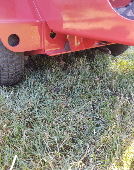The back of a red lawn mower sits on green grass, showing the frame, tires, and metal undercarriage.