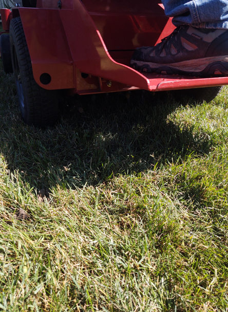 A low-angle view of a person's foot in a hiking shoe resting on the red metal foot platform of a lawn mower over grass.