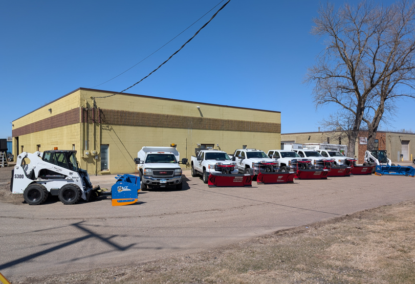 A fleet of trucks equipped with snow plows and a skid-steer loader parked in a lot outside a yellow commercial building.