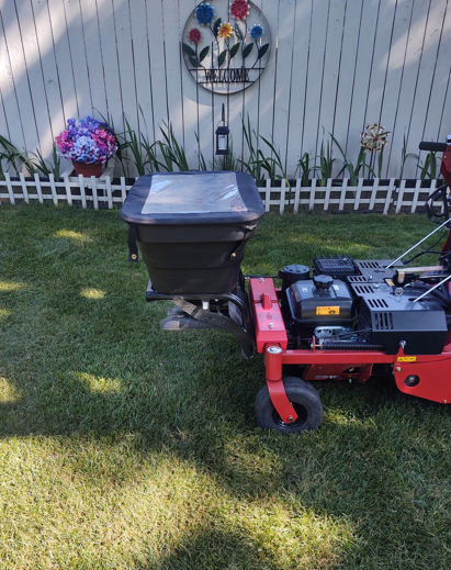 A red lawn mower with a black hopper attachment sits on a grassy lawn in front of a white picket fence.
