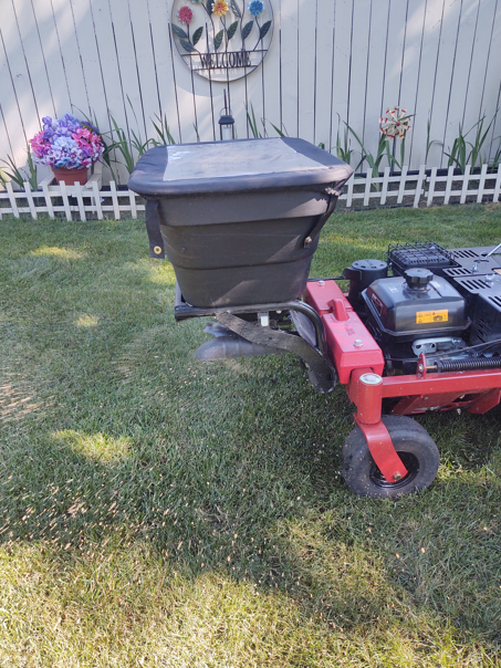 A black broadcast spreader mounted to the rear of a red lawn mower, situated on a residential lawn by a white picket fence.