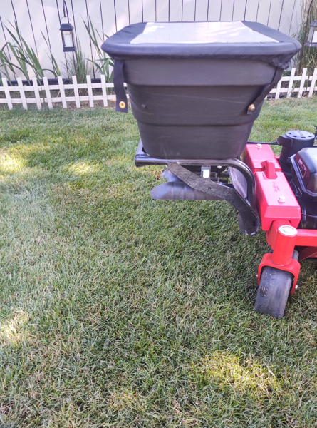 A black broadcast spreader mounted on a red lawn mower sits on a green lawn in front of a white fence.