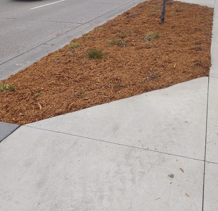 A landscaped triangular bed covered in brown wood mulch adjacent to a concrete sidewalk and asphalt road.