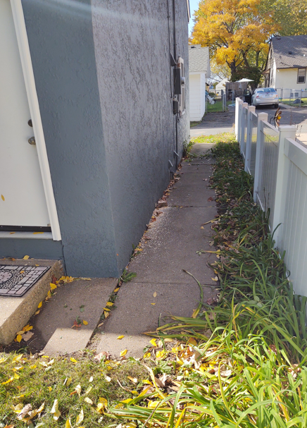 A narrow concrete walkway between a grey house wall and a white fence, scattered with fallen autumn leaves.