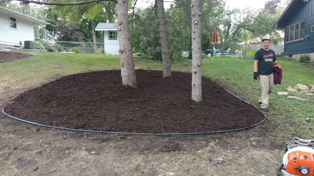 A person stands near a large, newly mulched tree bed outlined with black edging in a residential yard.