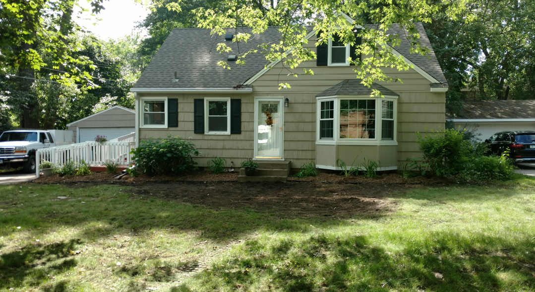 A tan, two-story house with a bay window, white trim, dark shutters, and a white fence in a lush, green, tree-lined yard.