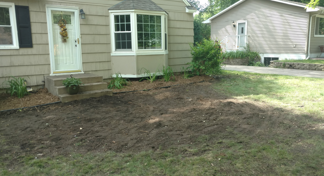 A light-colored house with a bay window and front door sits behind a patch of freshly tilled dark brown soil.
