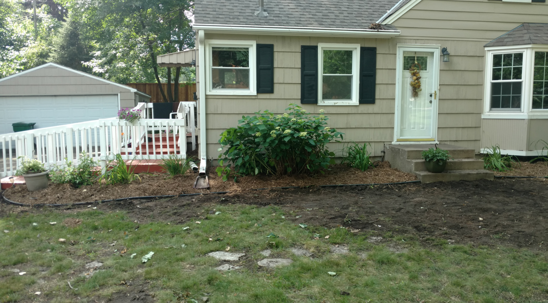 A tan house with a white front porch railing and a fresh mulch garden bed in front, with a detached garage to the left.