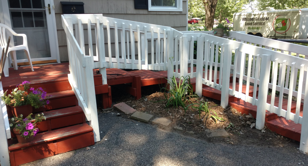 A red wooden ramp with white railings leads to a house entrance next to a small set of matching steps.