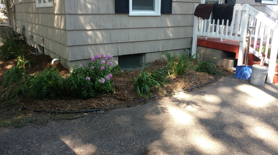 A light-colored house exterior with a small flower bed in front of a basement window and a white porch railing to the right.