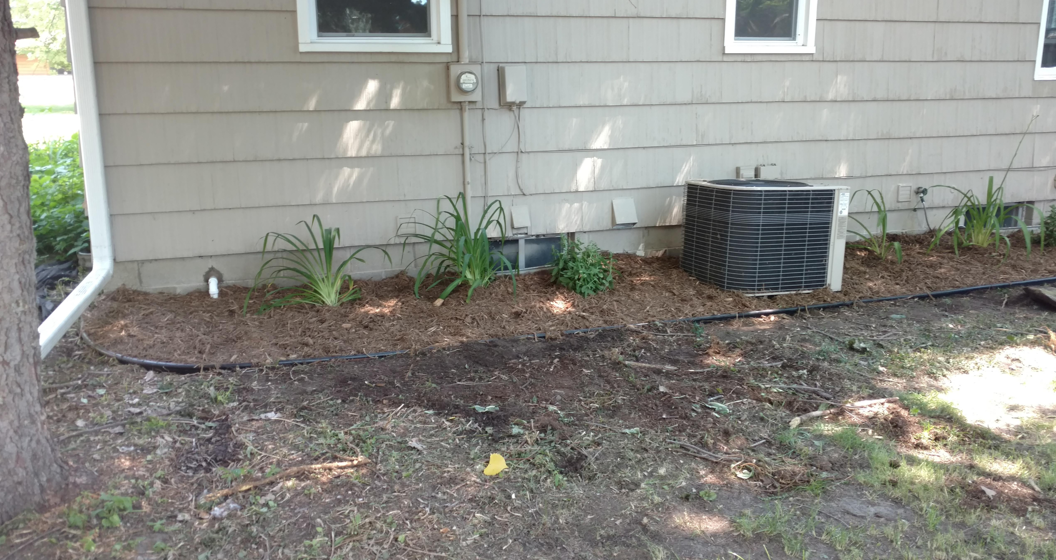 A tan-sided house with a mulch garden bed, several small green plants, and a central air conditioning unit.