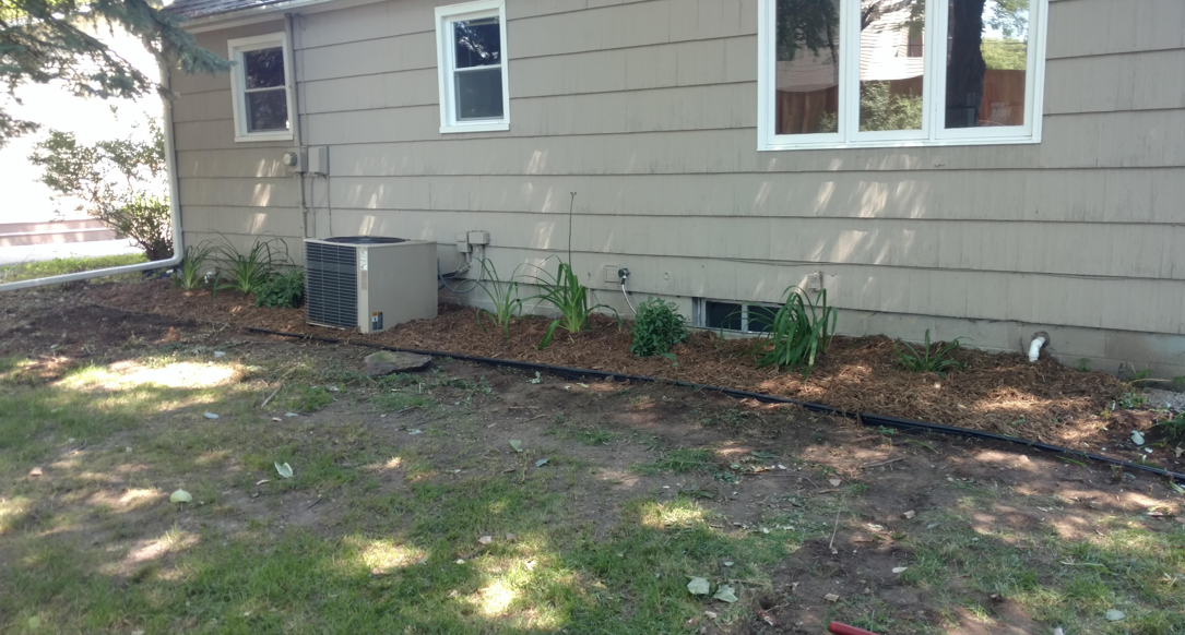 A side view of a house with tan siding, a window, an air conditioning unit, and a small flower bed with mulch.