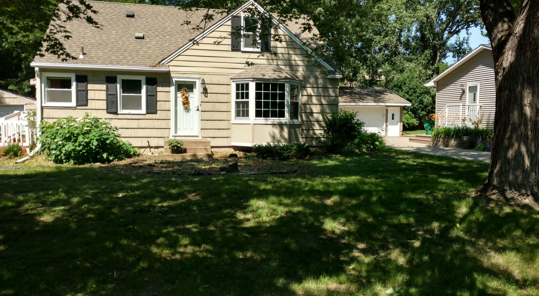 A tan-colored house with black shutters, a front bay window, and a detached garage in a leafy, sunlit residential yard.