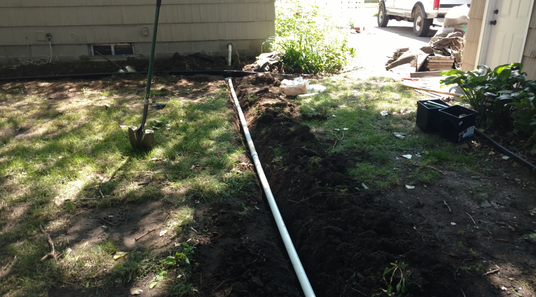 A white pipe lies inside a narrow trench dug through a grassy residential yard next to a house.