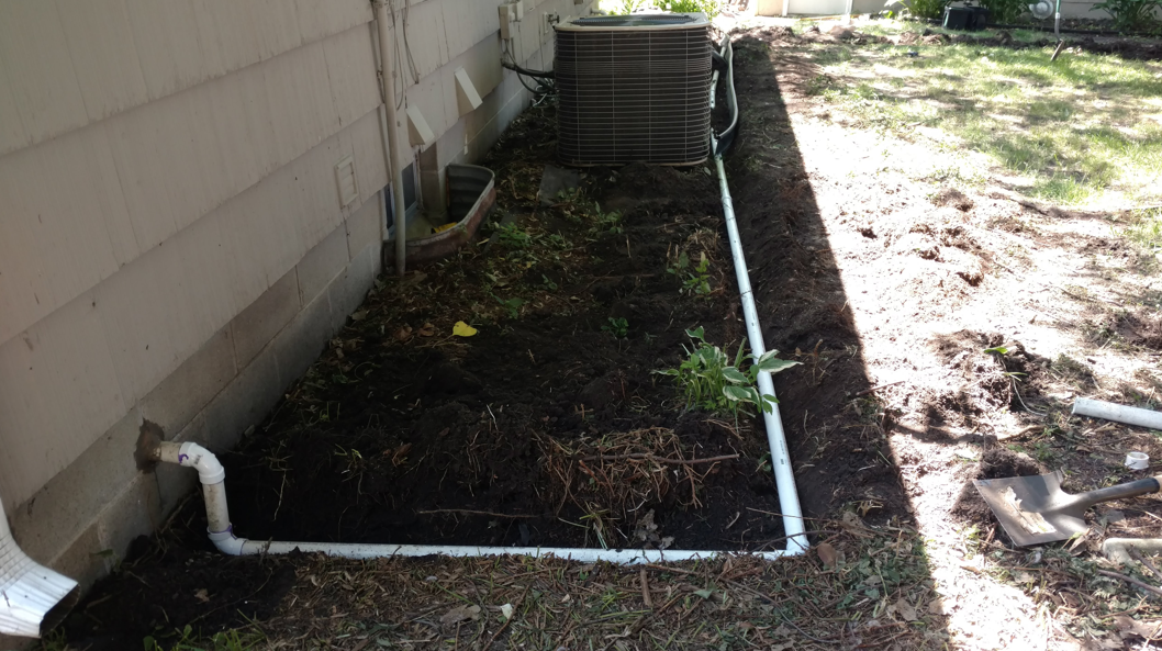 White PVC drainage pipes run along the side of a house, connecting a downspout to an area near an AC unit in a dirt bed.
