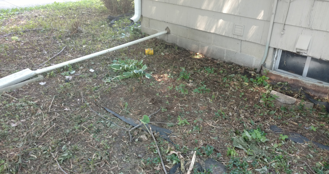 A white downspout extension lies on the ground next to the foundation of a beige house with a small basement window.