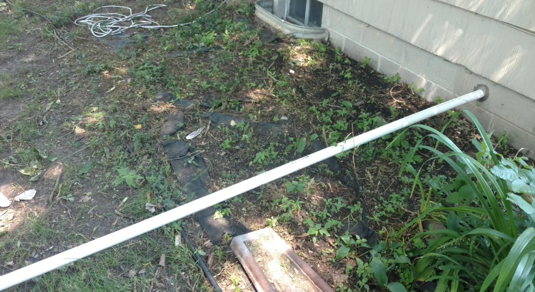 A white PVC pipe extending from the foundation of a house into a garden bed.