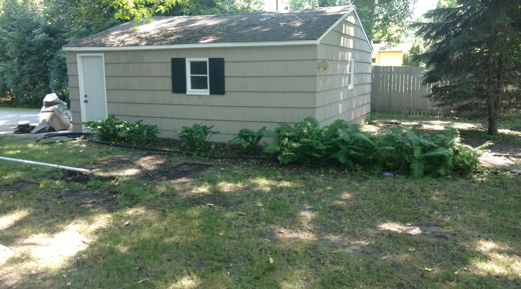 A light-tan shed with a white door, dark-shuttered window, and green plants in front on a grassy lawn.