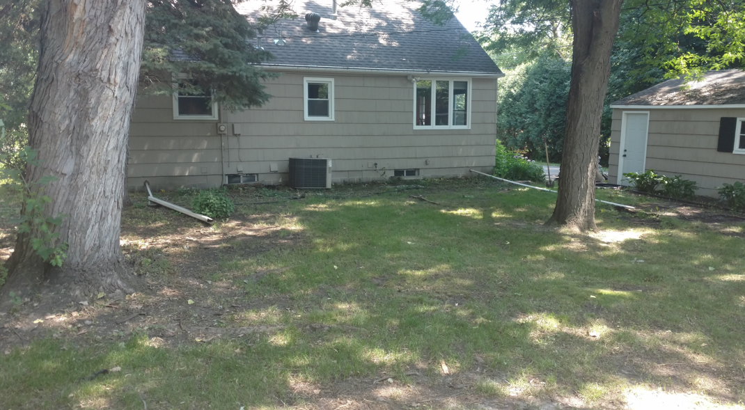 A tan house with a gray roof and a detached garage in a yard with large trees and patches of lawn.