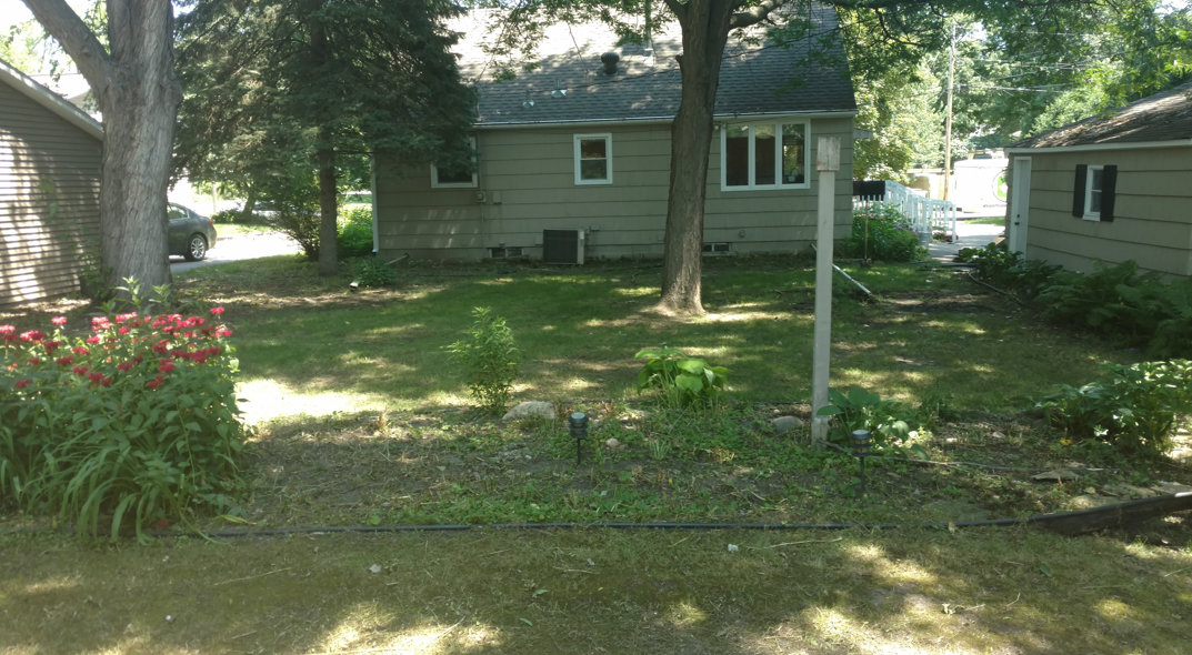 A backyard view featuring a light-colored house, a grassy lawn, mature trees, and a garden bed with red flowers.