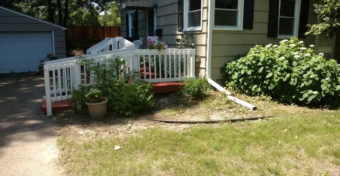A white wooden wheelchair ramp leading up to the front entrance of a house, surrounded by plants and a patch of grass.