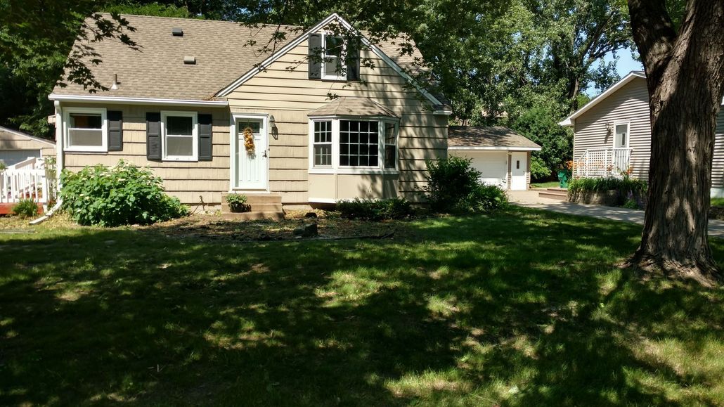 A one-story tan house with a white door, a bay window, a gabled roof, and a detached garage on a grassy, tree-filled lot.