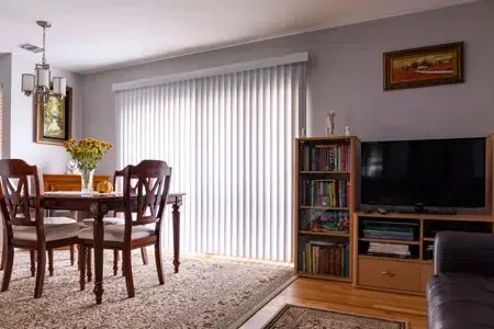 Bright living-dining room with wooden table, TV, bookshelf, and vertical blinds by sliding doors