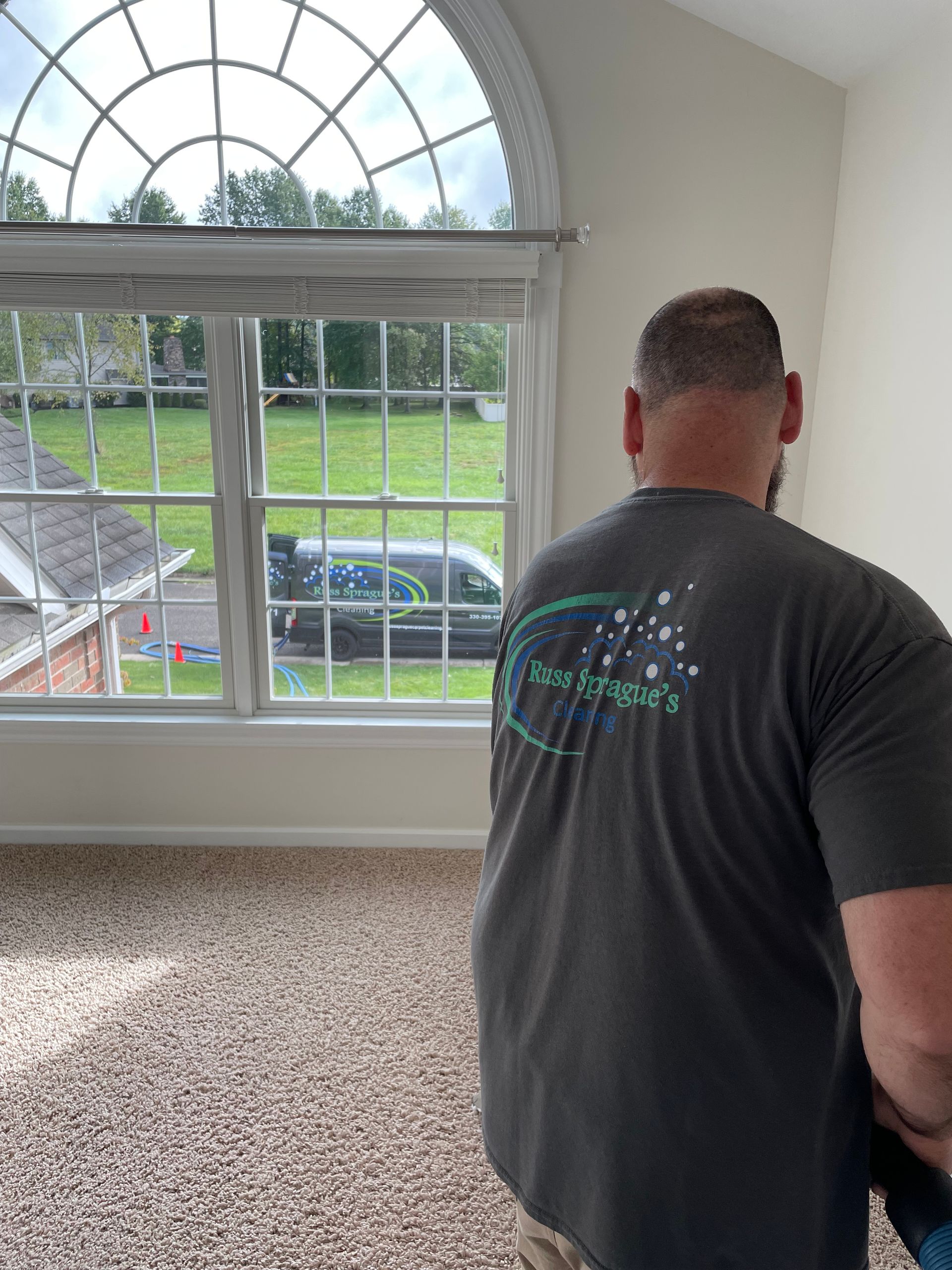 Man standing in a carpeted room, looking out a large arched window onto a green yard.