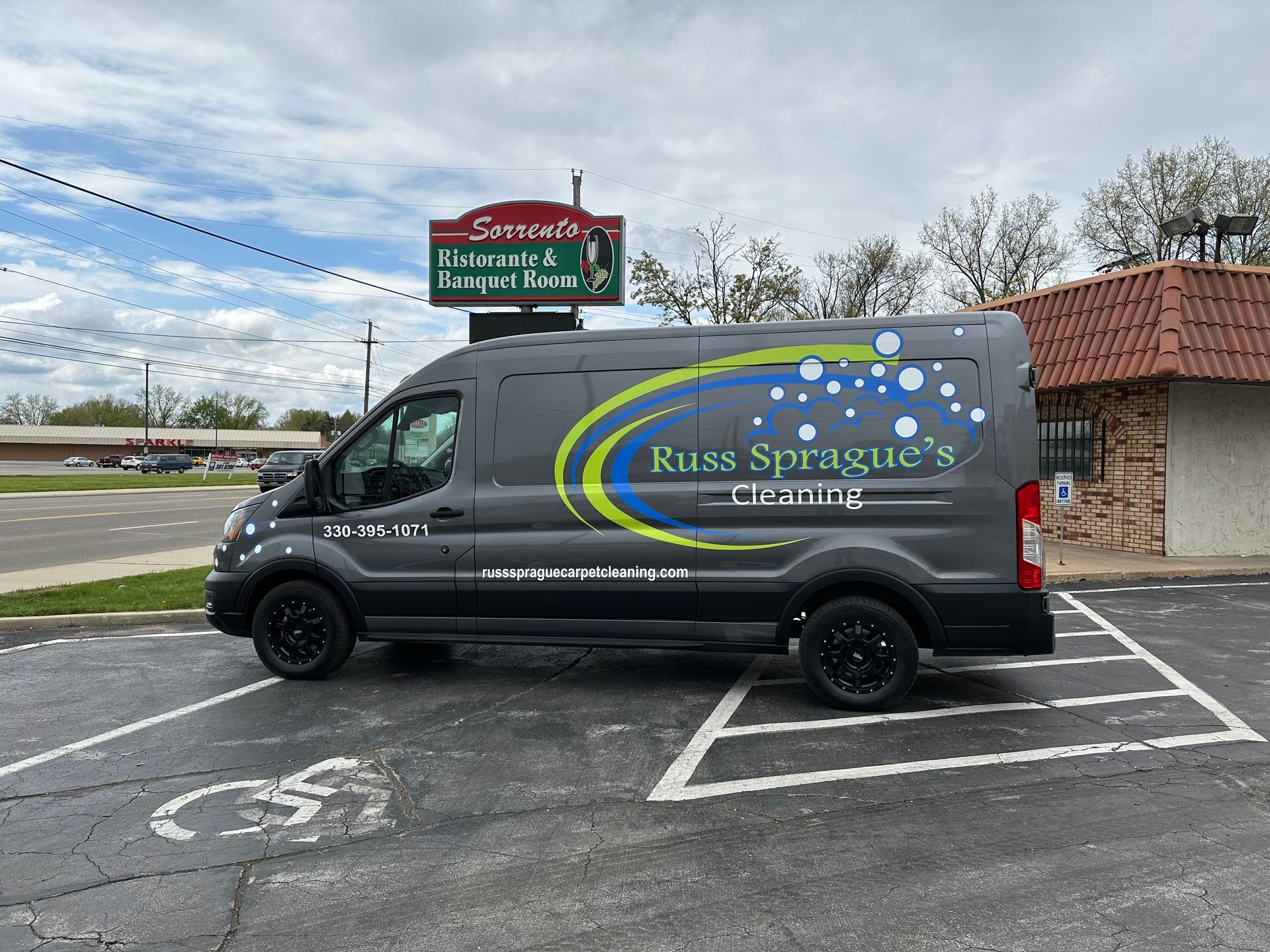 Dark gray service van with “Kiss Sprague” logo parked in a lot beside a brick building.