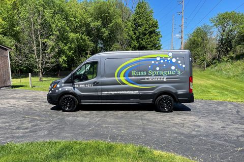 Gray service van parked on a driveway, with “Russ Services” logo on the side, trees in the background.