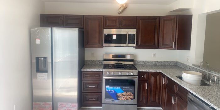 A kitchen with dark wood cabinets, stainless steel appliances, and gray countertops. A refrigerator stands next to a stove.