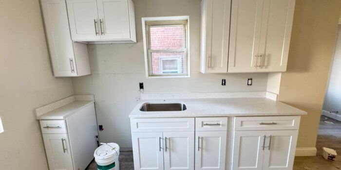 A small, white kitchen featuring cabinets and a countertop with a sink. A window is set in the center with construction visible.