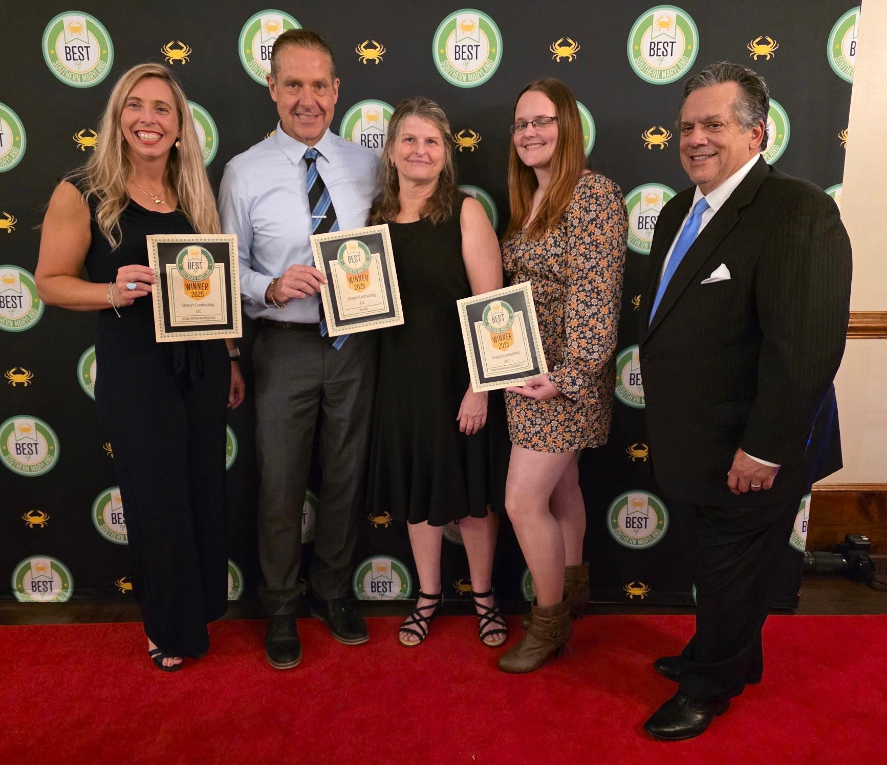 Five people holding framed awards on a red carpet with a themed backdrop.