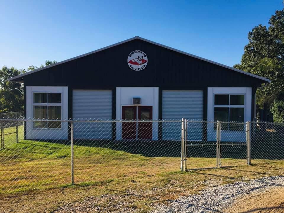 A large black building with a red door is behind a chain link fence.