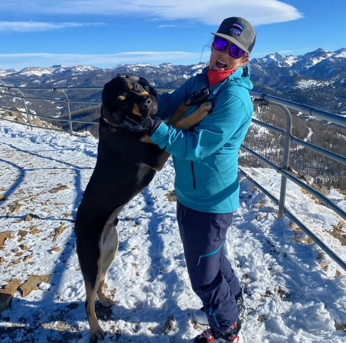 Woman in blue jacket hugs a large black dog on a snowy mountaintop with a mountain range in the background.