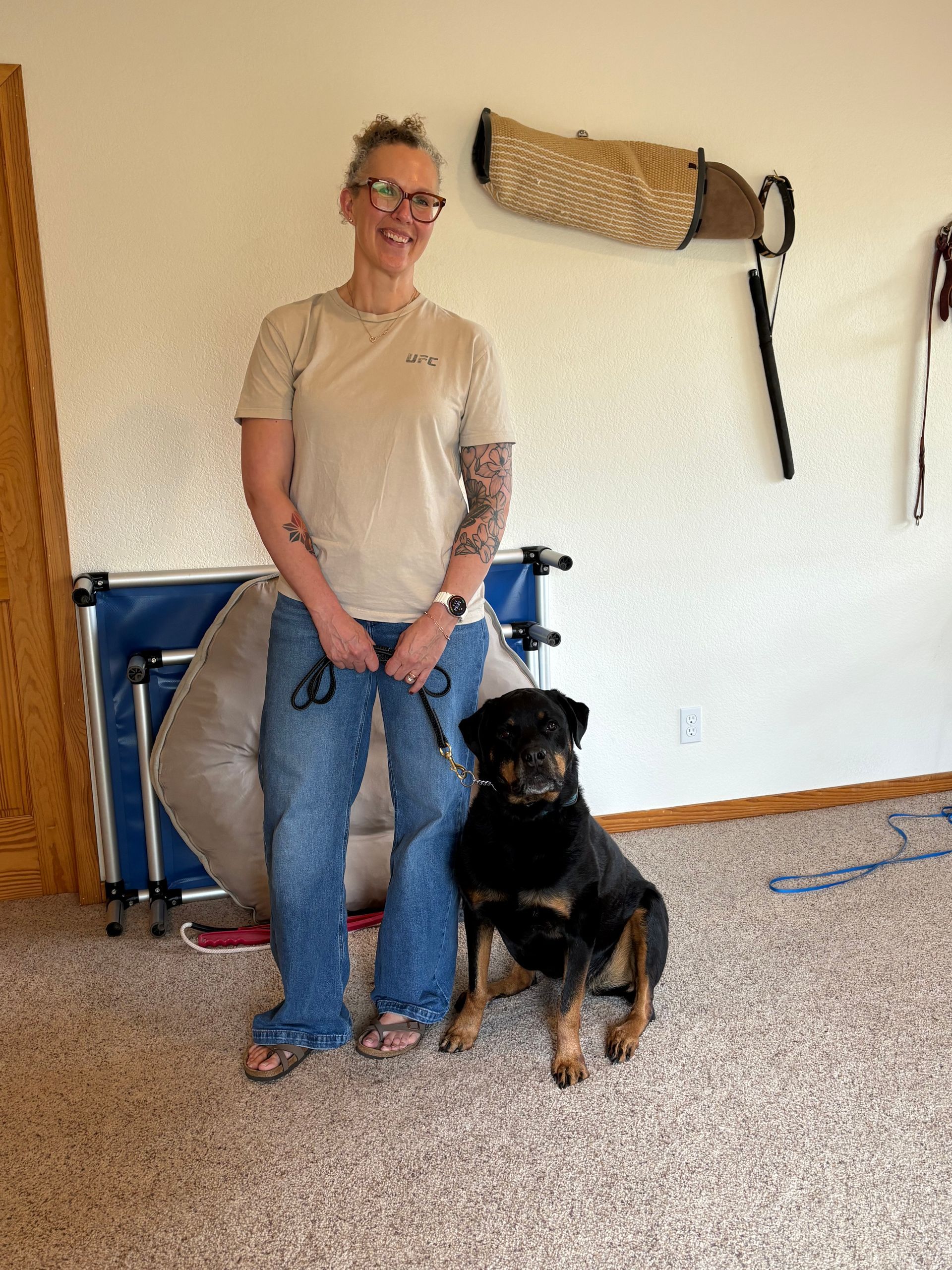 Woman and Rottweiler dog posing together in an indoor setting. Beige wall, training equipment.