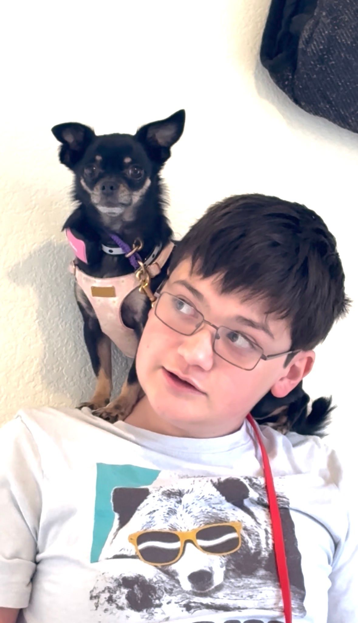 A person wearing glasses smiles with a small black dog on their shoulder; both look towards the viewer.