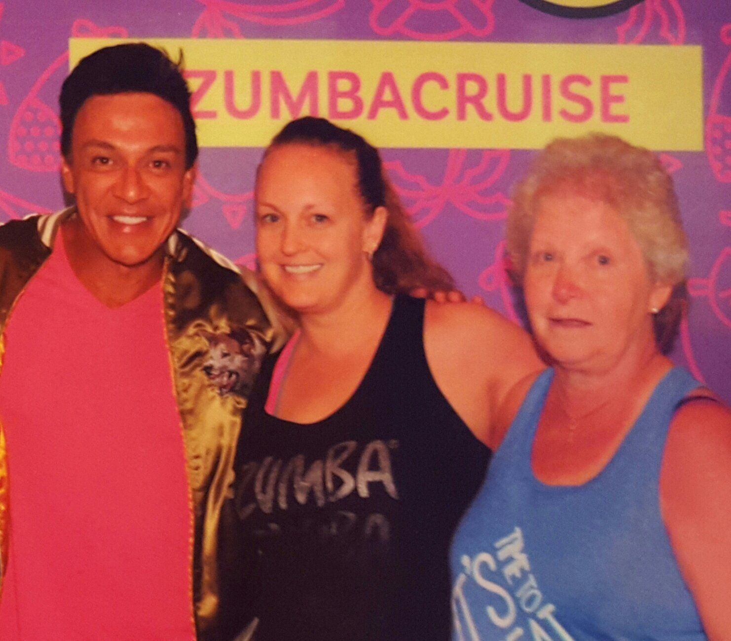 A man and two women pose for a photo in front of a Zumba Cruise sign.